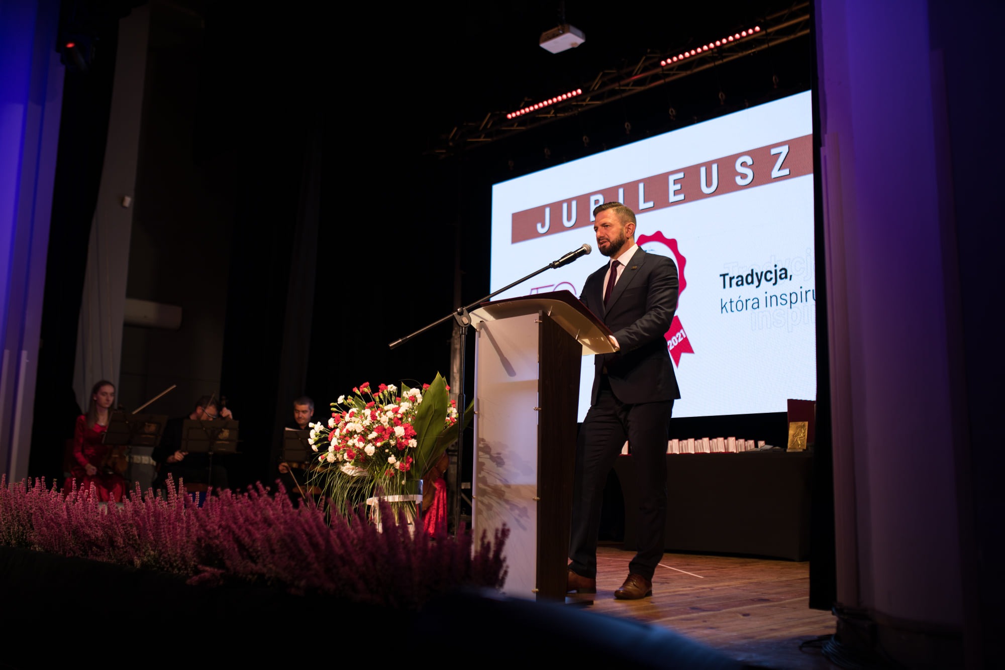 Prelegent w ciemnym garniturze przemawia na scenie podczas jubileuszu, za nim ekran z napisem 'Jubileusz' i hasłem 'Tradycja, która inspiruje', a po lewej stronie orkiestra smyczkowa.