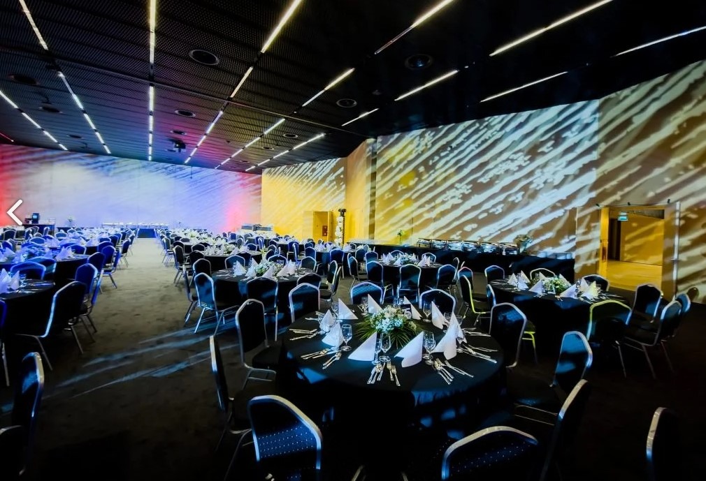 Sala bankietowa z okrągłymi stołami nakrytymi czarnymi obrusami, gotowa na przyjęcie. Dekoracje kwiatowe na stołach, oświetlenie sceniczne w tle.