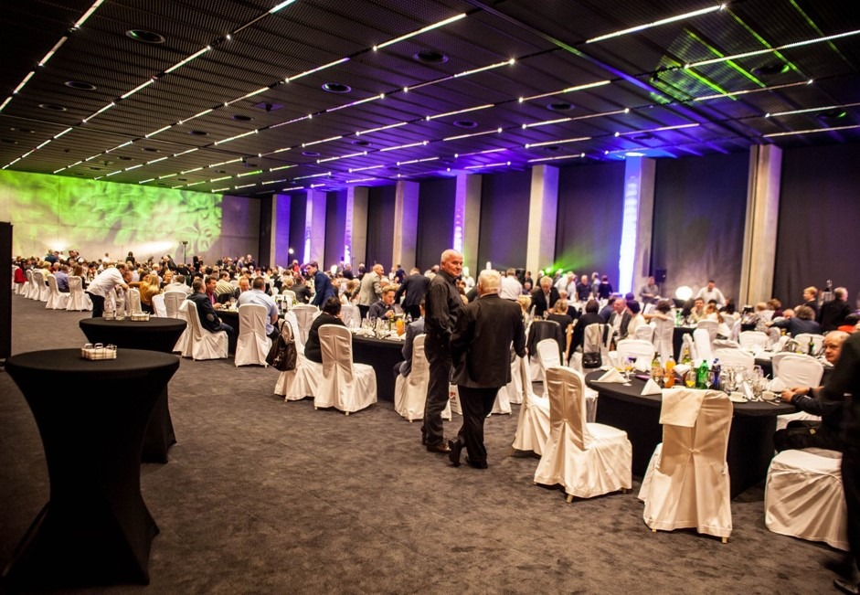 Sala bankietowa z gośćmi siedzącymi przy okrągłych stołach, oświetlona kolorowymi światłami. Widoczne kolumny i dekoracje. Uczestnicy w strojach formalnych.
