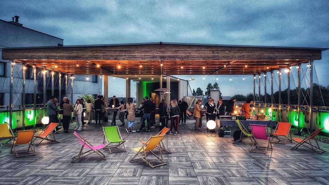 Impreza firmowa na tarasie z drewnianą konstrukcją, oświetleniem i kolorowymi leżakami. Ludzie w tle, wieczorny klimat.