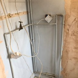 DR.HOUSE - Fragment wnętrza w trakcie remontu: odsłonięte przewody elektryczne, rury i surowa ściana z resztkami starej farby. Widoczny etap prac instalacyjnych i przygotowawczych.