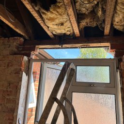 DR.HOUSE - Demontaż okna: widok na ceglaną ścianę z wyciętym otworem okiennym, izolacja stropu i drabina oparta o ramę okienną. Prace remontowe w toku.