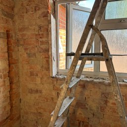 DR.HOUSE - Odsłonięta cegła w trakcie renowacji przy oknie, z metalową drabiną opartą o ścianę. Widoczne ślady po starym tynku i gruz na podłodze. Intensywne światło dzienne.