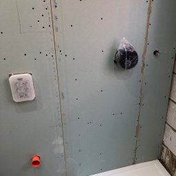 DR.HOUSE - Ściana z płyty karton-gips w łazience w trakcie remontu, widoczne otwory i elementy instalacji, w tym rura i puszka pod baterię. Fragment brodzika w dolnym rogu.