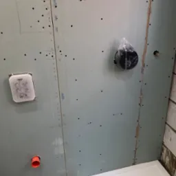 Ściana z płyty karton-gips w łazience w trakcie remontu, widoczne otwory i elementy instalacji, w tym rura i puszka pod baterię. Fragment brodzika w dolnym rogu.