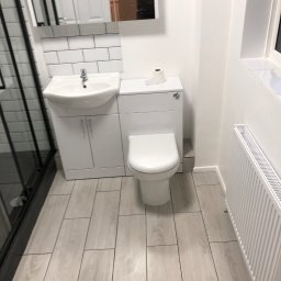 DR.HOUSE - Nowoczesna, jasna łazienka po remoncie: białe płytki, umywalka z szafką, toaleta, grzejnik. W lustrze odbija się osoba robiąca zdjęcie.