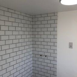 DR.HOUSE - Białe, prostokątne płytki z czarną fugą ułożone na ścianach w rogu łazienki. Widoczne wystające rury i fragment podłogi z ciemnymi płytkami. Górne oświetlenie.