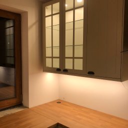 DR.HOUSE - Kuchenny narożnik z drewnianym blatem i szarymi szafkami wiszącymi z przeszklonymi frontami, podświetlonymi od dołu. Widoczne okno w tle.