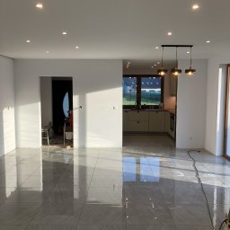 DR.HOUSE - Jasne, przestronne wnętrze z nową, błyszczącą szarą podłogą z płytek. Widok na kuchnię i drzwi przesuwne. Świeżo po remoncie, gotowe do umeblowania.