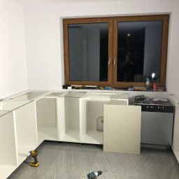 DR.HOUSE - Montaż białych szafek kuchennych pod oknem z drewnianą ramą. Na podłodze szare płytki i narzędzia: wiertarki, ściski stolarskie. Widoczny fragment zabudowy zmywarki.