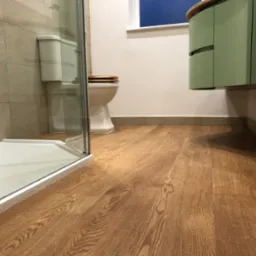 Wykończona łazienka z drewnianą podłogą, kabiną prysznicową i umywalką w odcieniach zieleni, z toaletą w tle. Ujęcie z niskiego kąta.