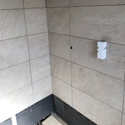 DR.HOUSE - Wykończony narożnik łazienki z beżowymi płytkami, widoczny fragment podłogi z czarną izolacją. Na ścianie biały element instalacyjny.