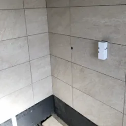 Wykończony narożnik łazienki z beżowymi płytkami, widoczny fragment podłogi z czarną izolacją. Na ścianie biały element instalacyjny.