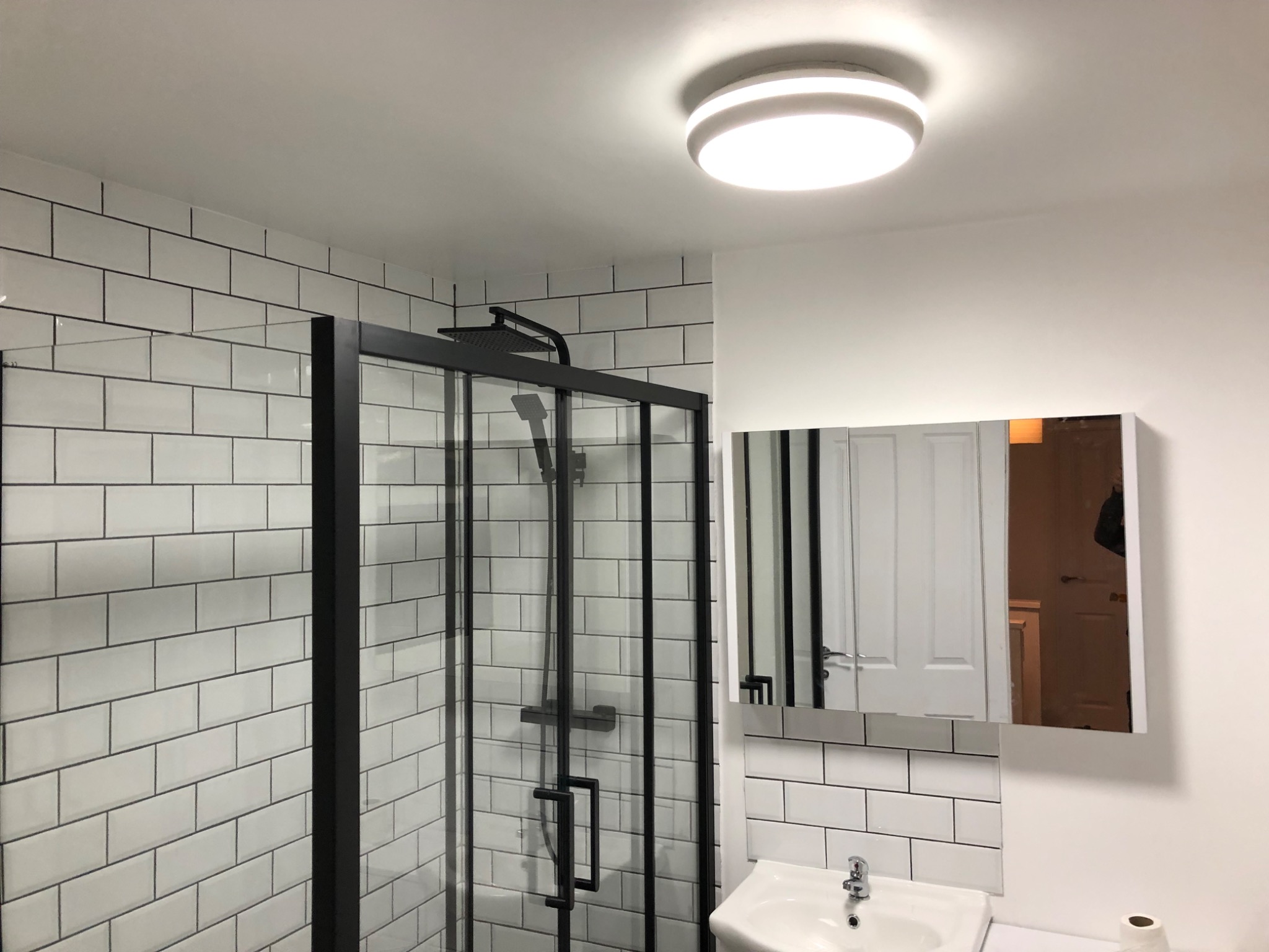 Nowoczesna kabina prysznicowa z czarną armaturą i białymi, prostokątnymi płytkami. W tle lustro nad umywalką, oświetlenie sufitowe.