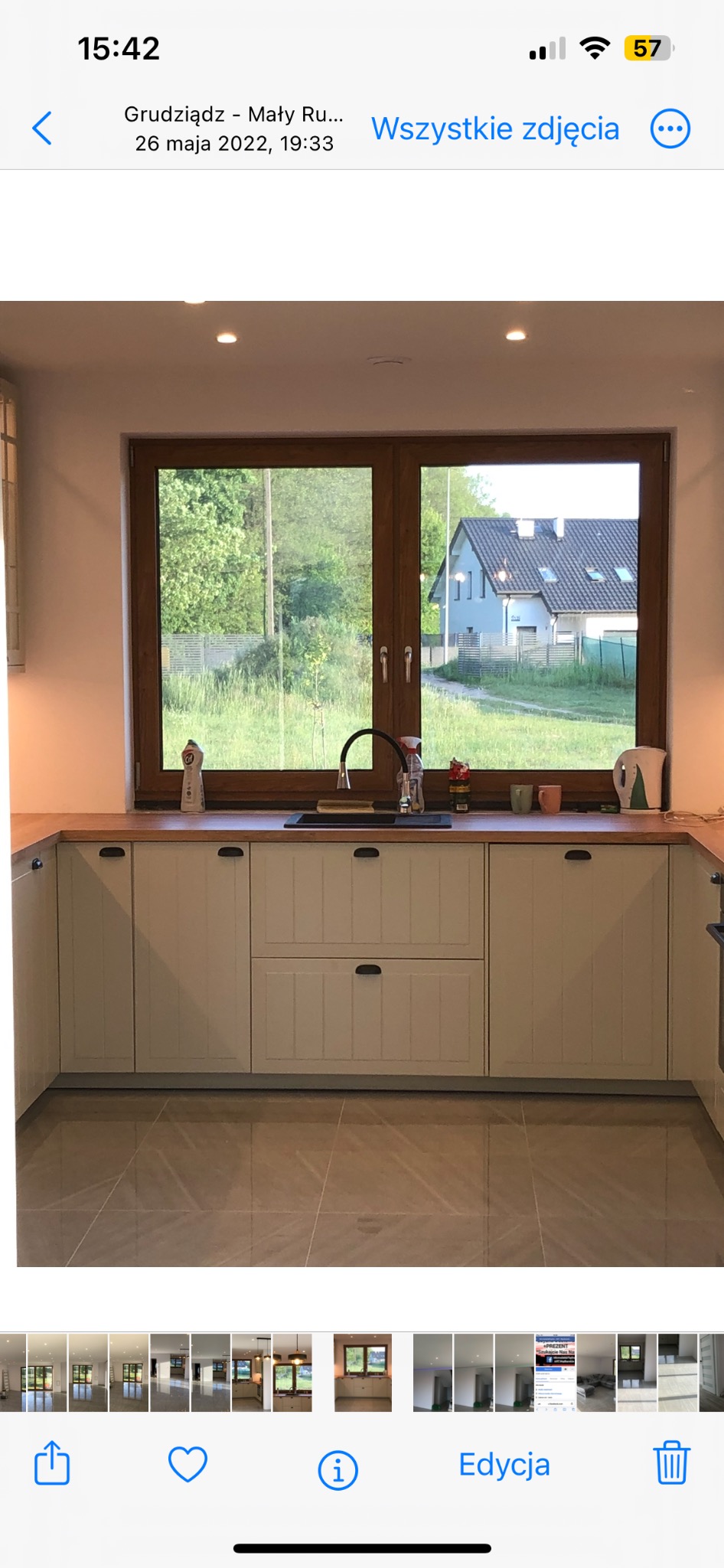 Jasna kuchnia z drewnianym blatem i widokiem na zieleń za oknem. Fronty szafek w stylu rustykalnym, beżowe płytki podłogowe. Umywalka pod oknem.