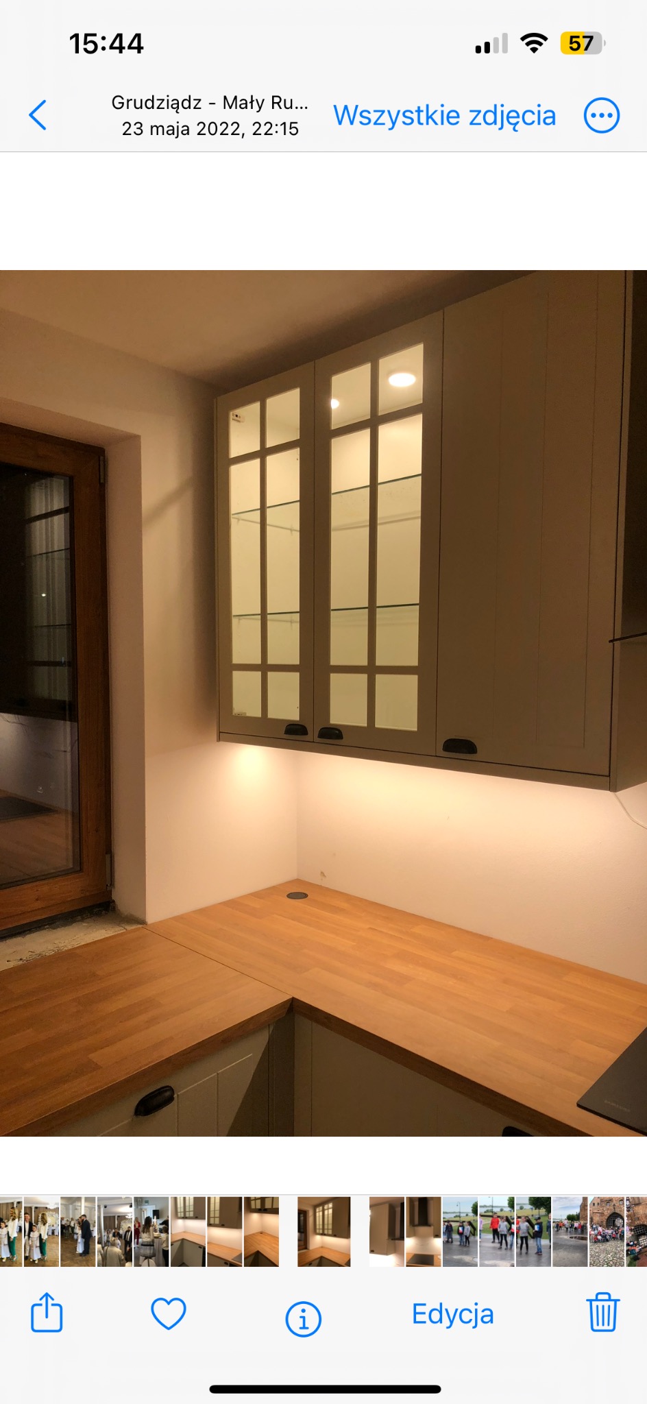 Kuchenny narożnik z drewnianym blatem i szarymi szafkami wiszącymi z przeszklonymi frontami, podświetlonymi od dołu. Widoczne okno w tle.