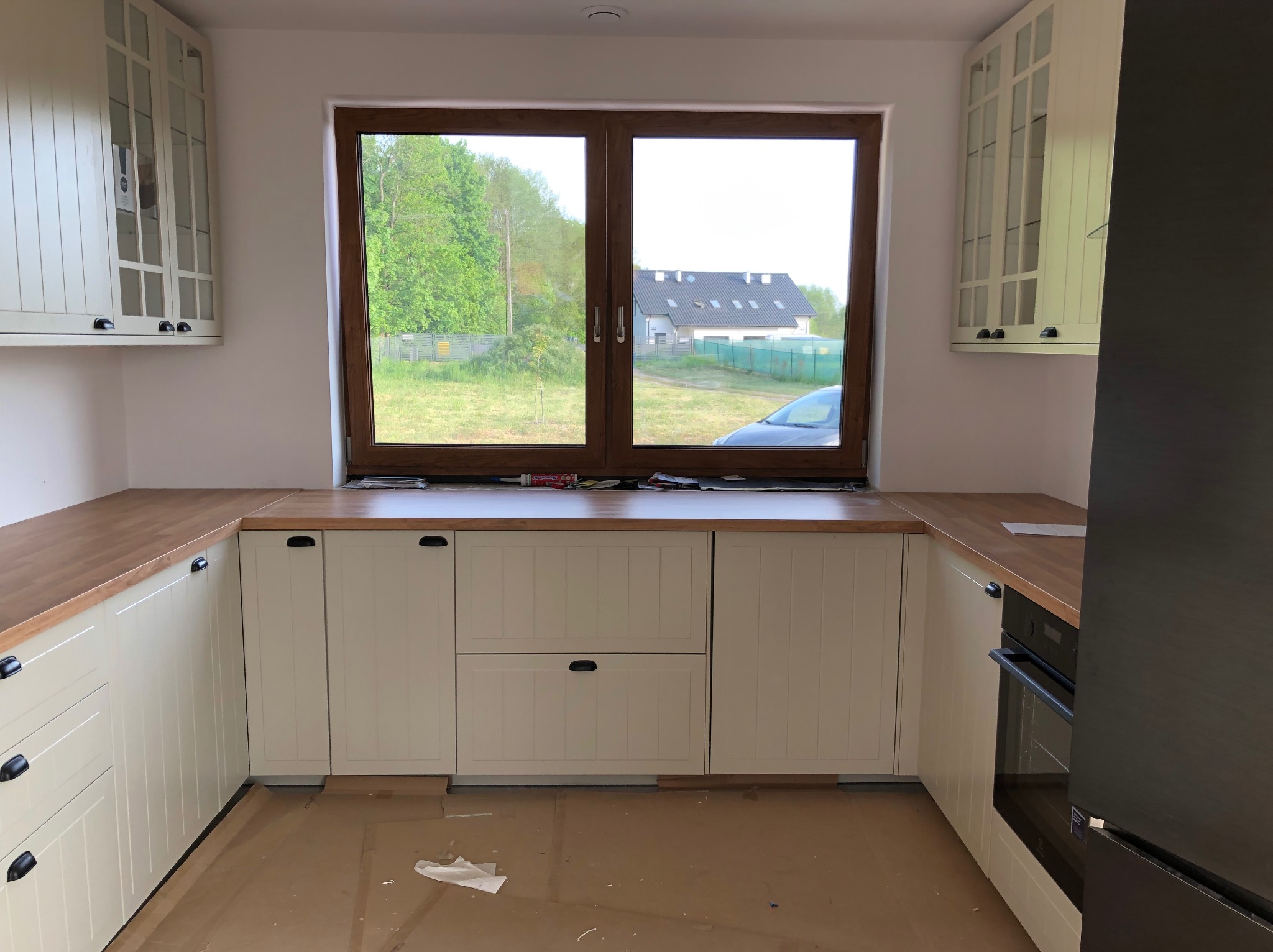 Jasna kuchnia z drewnianym blatem, okno z widokiem na zieleń. Zabudowa kuchenna z frezowanymi frontami i czarnymi uchwytami. Nowoczesny piekarnik w zabudowie.