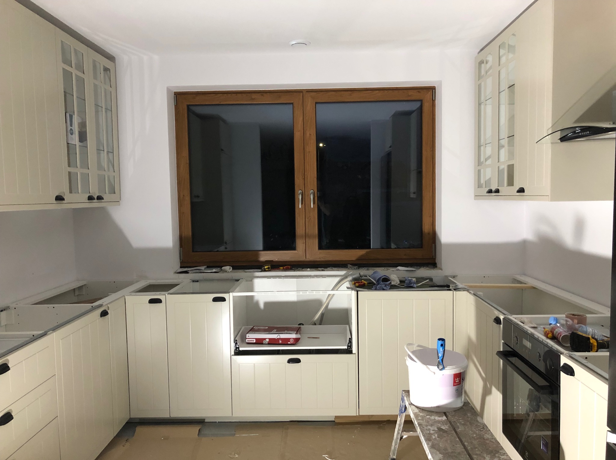Jasna kuchnia w trakcie montażu: kremowe szafki, drewniane okno. Widoczne narzędzia i drabina malarska. Etap prac wykończeniowych.