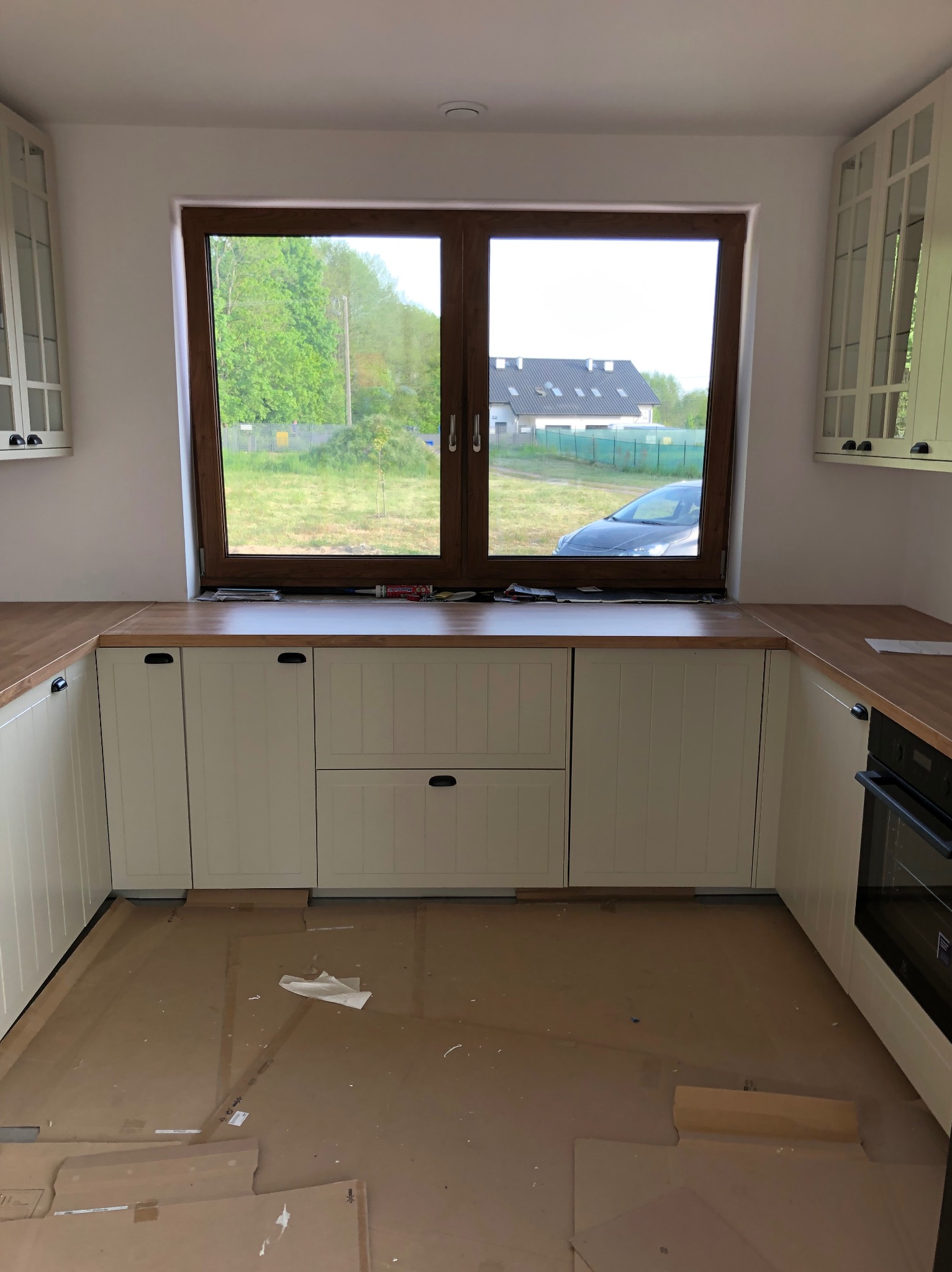 Jasna kuchnia w trakcie montażu: białe szafki z drewnianym blatem pod oknem z widokiem na zieleń. Na podłodze kartony zabezpieczające.