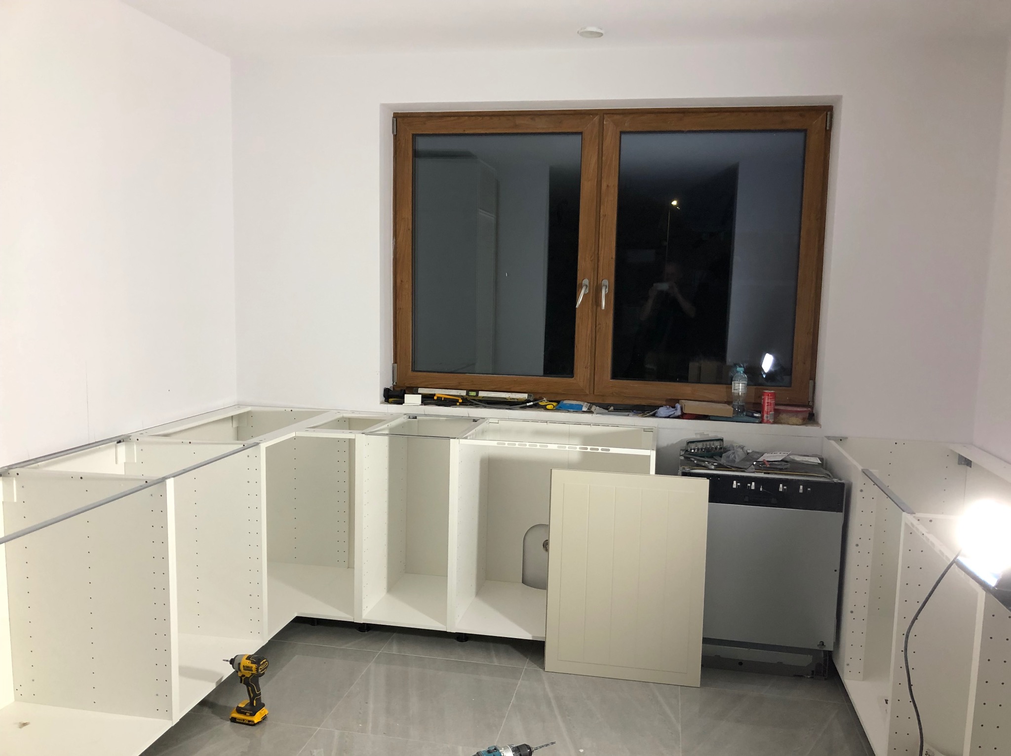 Montaż białych szafek kuchennych pod oknem z drewnianą ramą. Na podłodze szare płytki. Widoczne narzędzia: wkrętarki. Etap prac: instalacja korpusów szafek.