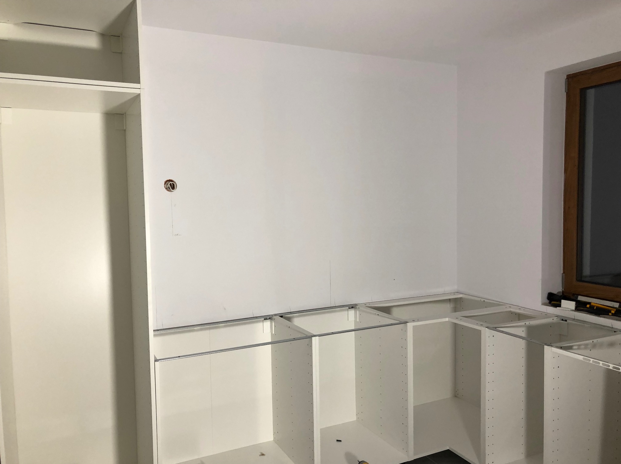 Montaż białych szafek kuchennych narożnych, widoczne prowadnice i szyny montażowe. Świeżo pomalowana ściana z gniazdkiem elektrycznym i drewniane okno w tle.