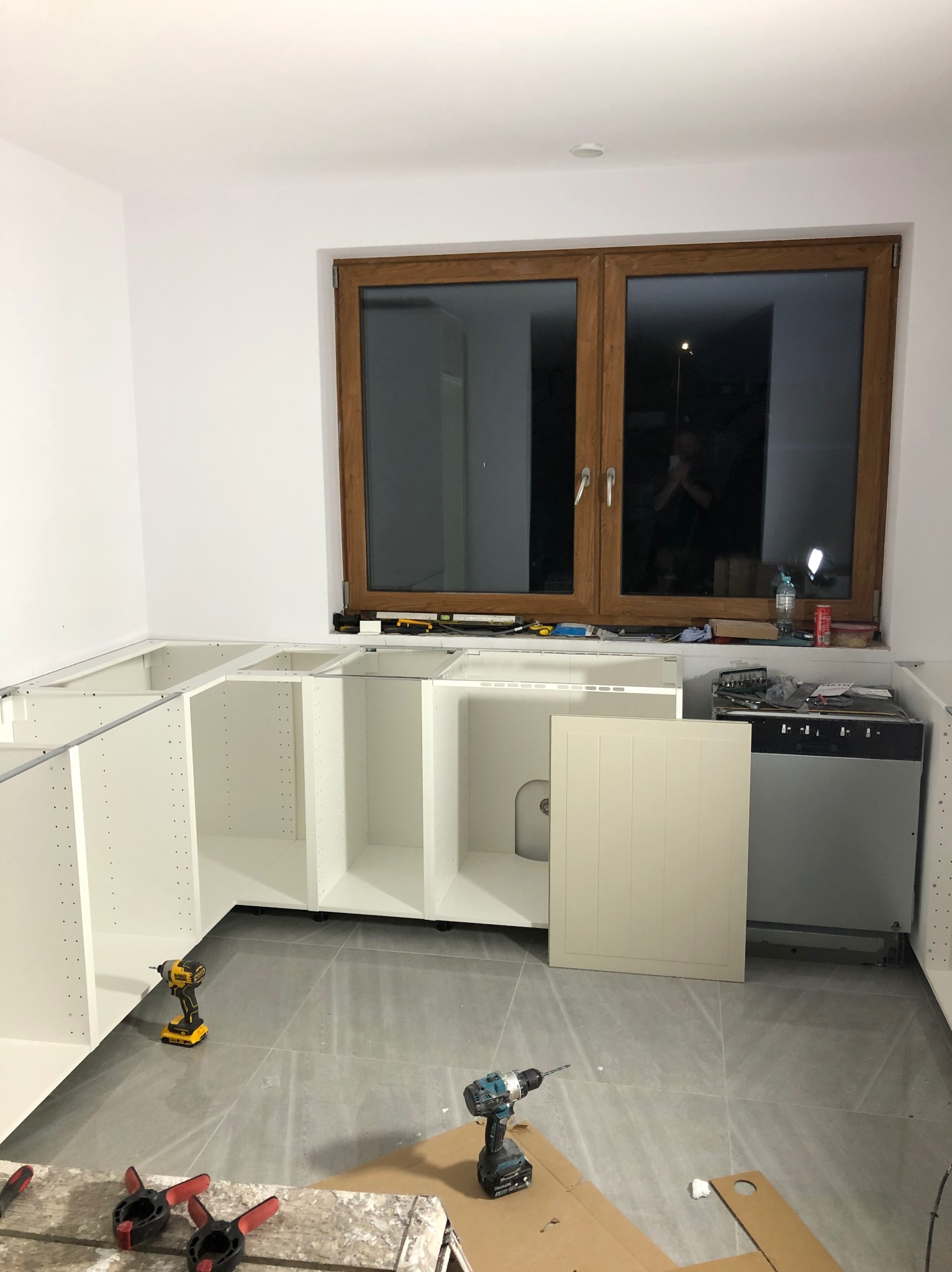 Montaż białych szafek kuchennych pod oknem z drewnianą ramą. Na podłodze szare płytki i narzędzia: wiertarki, ściski stolarskie. Widoczny fragment zabudowy zmywarki.
