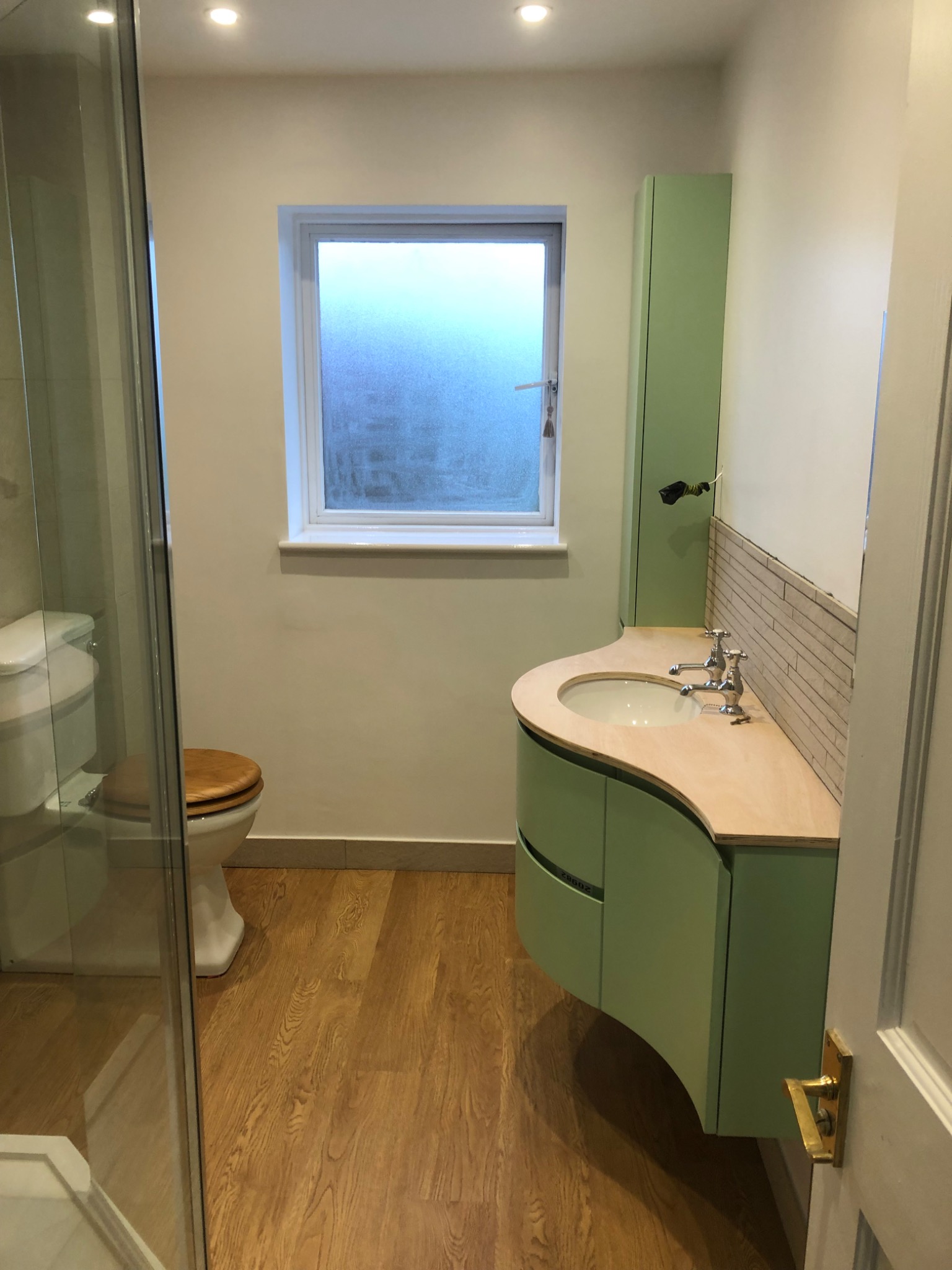 Jasna łazienka po remoncie: kabina prysznicowa, toaleta z drewnianą deską, umywalka z szafką w odcieniach zieleni, drewnopodobna podłoga i okno z widokiem na zewnątrz.