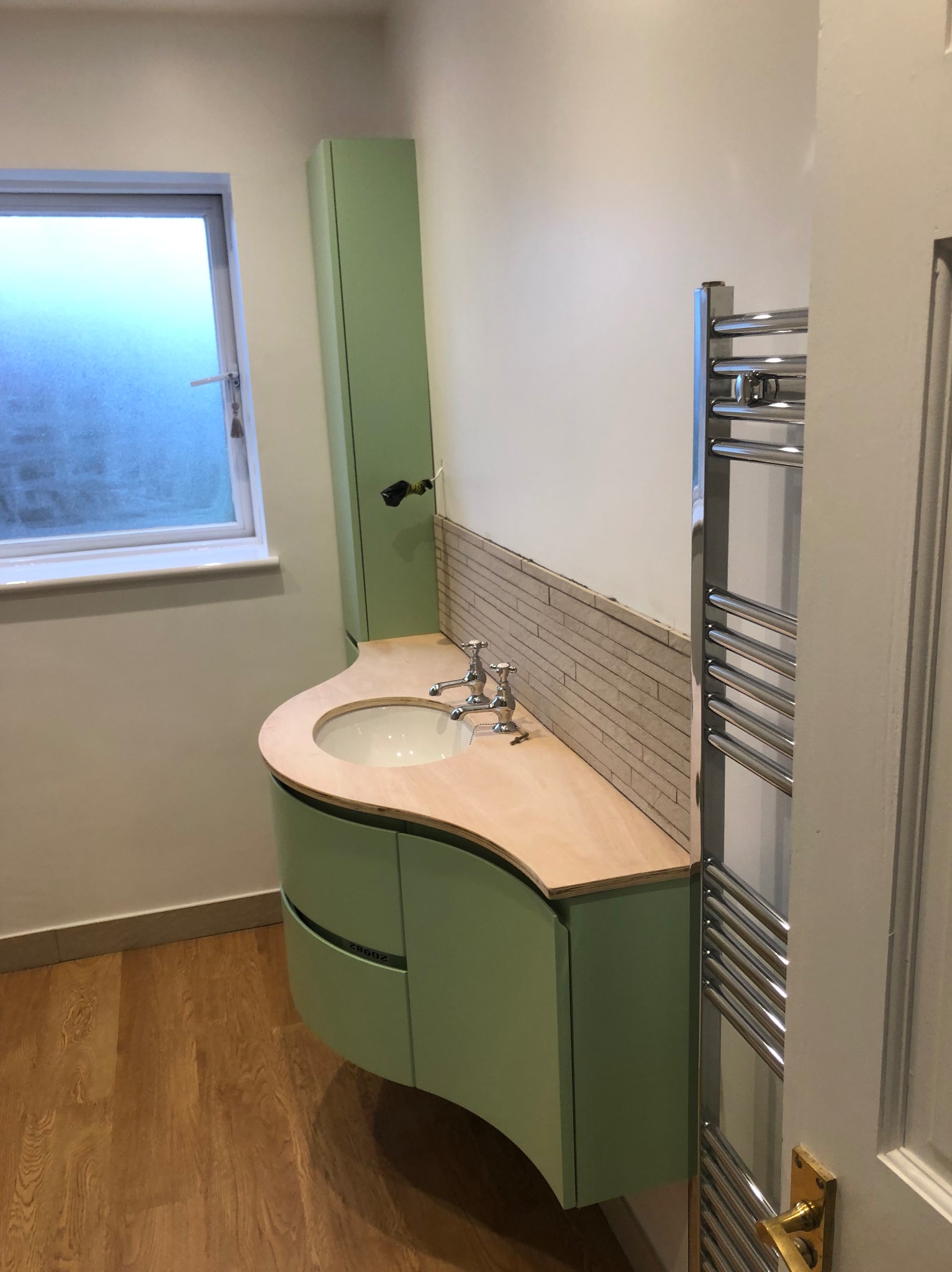 Wykończona łazienka z umywalką w pastelowej zieleni, drewnianym blatem i mozaiką. Grzejnik drabinkowy obok drzwi. Widok na okno i drewnianą podłogę.