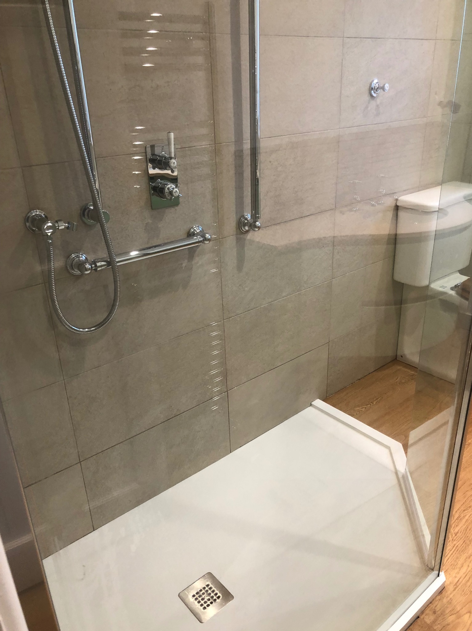 Nowoczesny prysznic z beżowymi płytkami, chromowaną armaturą i szklaną kabiną. Widoczny odpływ liniowy i fragment toalety. Jasne, minimalistyczne wykończenie łazienki.