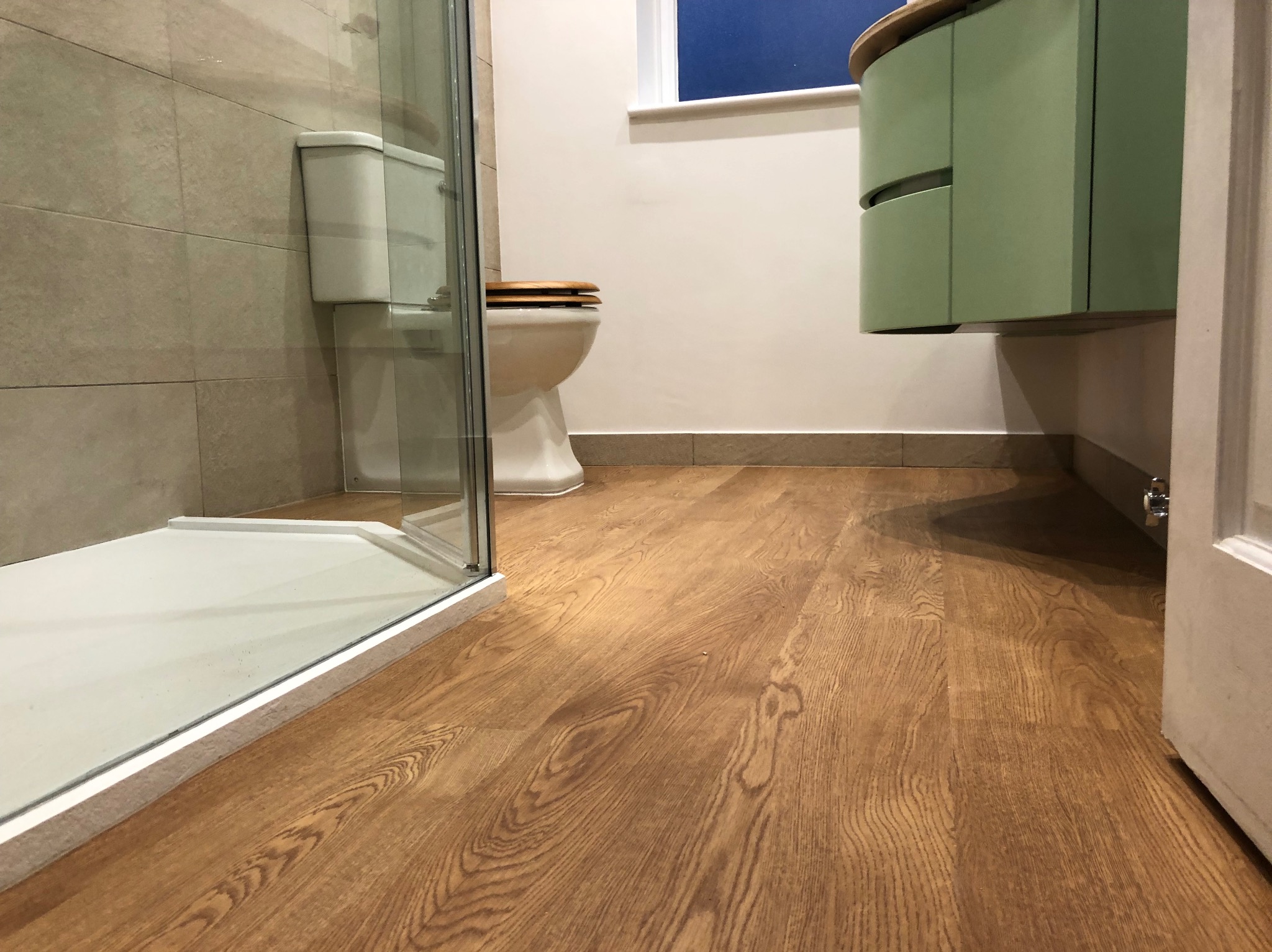 Wykończona łazienka z drewnianą podłogą, kabiną prysznicową i umywalką w odcieniach zieleni, z toaletą w tle. Ujęcie z niskiego kąta.