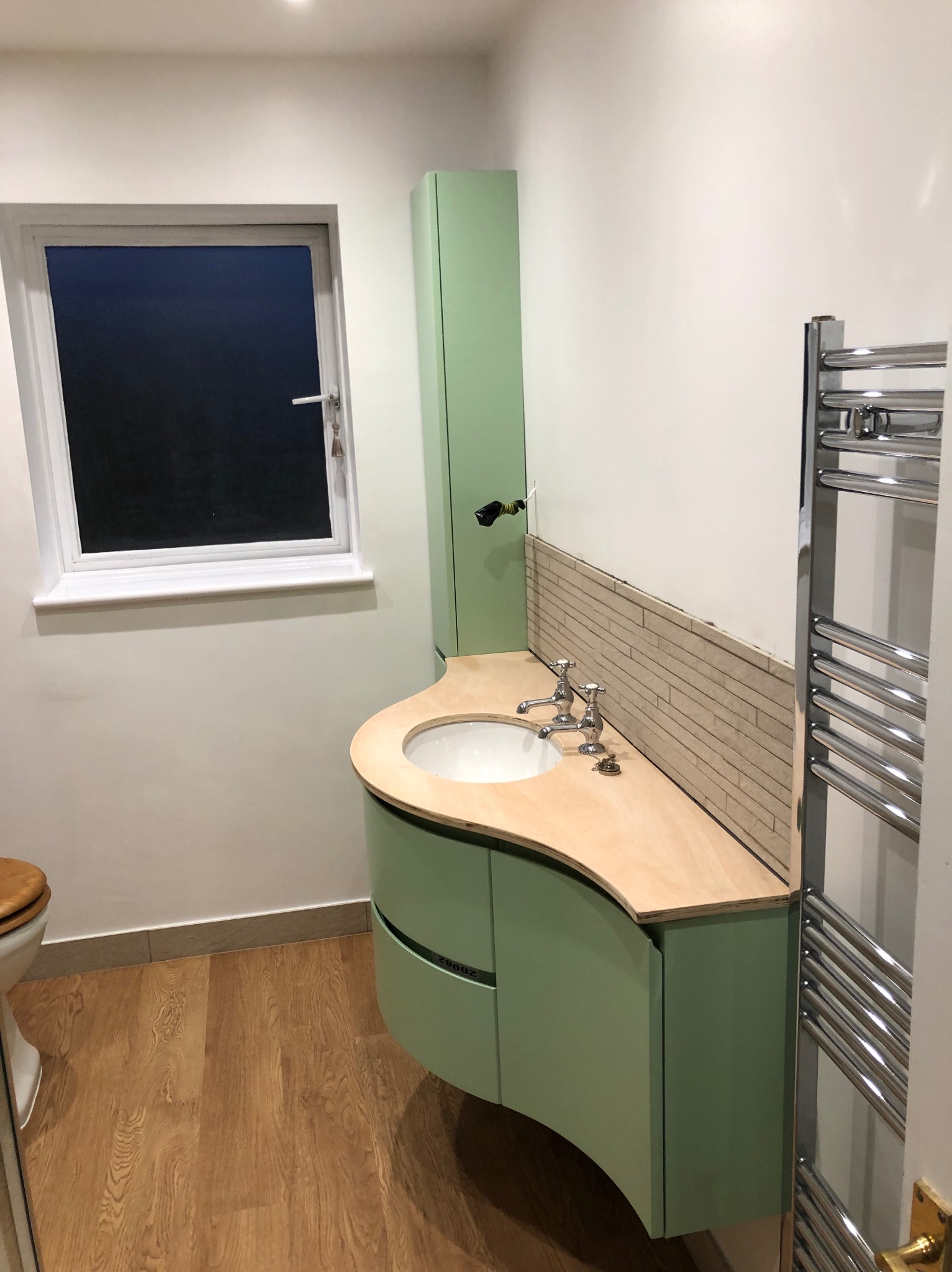 Wykończona łazienka w Grudziądzu: zielona szafka z umywalką, drewniany blat, częściowo ułożone płytki, grzejnik drabinkowy, okno z widokiem na zewnątrz.