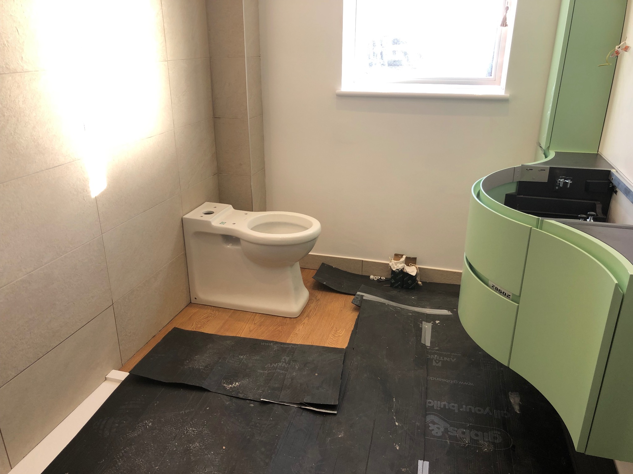 Montaż toalety i umywalki w łazience z beżowymi płytkami i drewnianą podłogą zabezpieczoną folią. Umywalka w odcieniach zieleni.