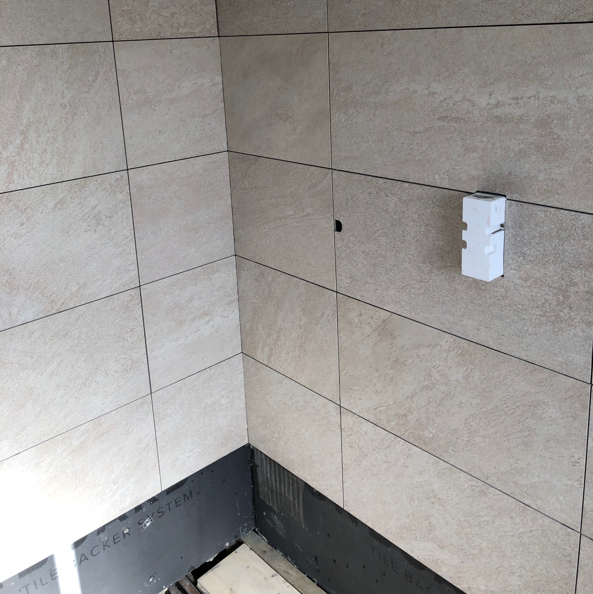 Wykończony narożnik łazienki z beżowymi płytkami, widoczny fragment podłogi z czarną izolacją. Na ścianie biały element instalacyjny.