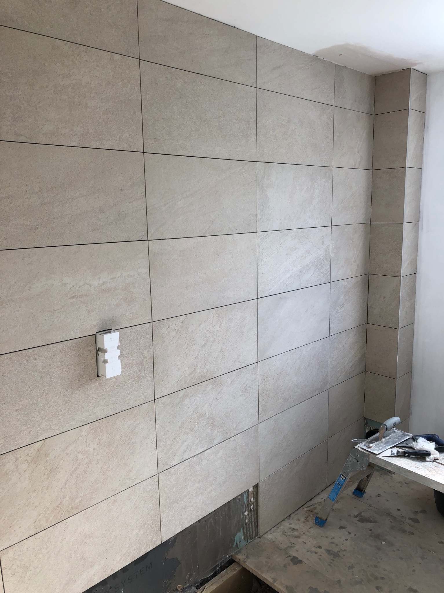 Ściana z beżowych płytek w łazience w trakcie remontu. Widoczna puszka elektryczna i drabina z narzędziami. Fragment podłogi bez płytek.