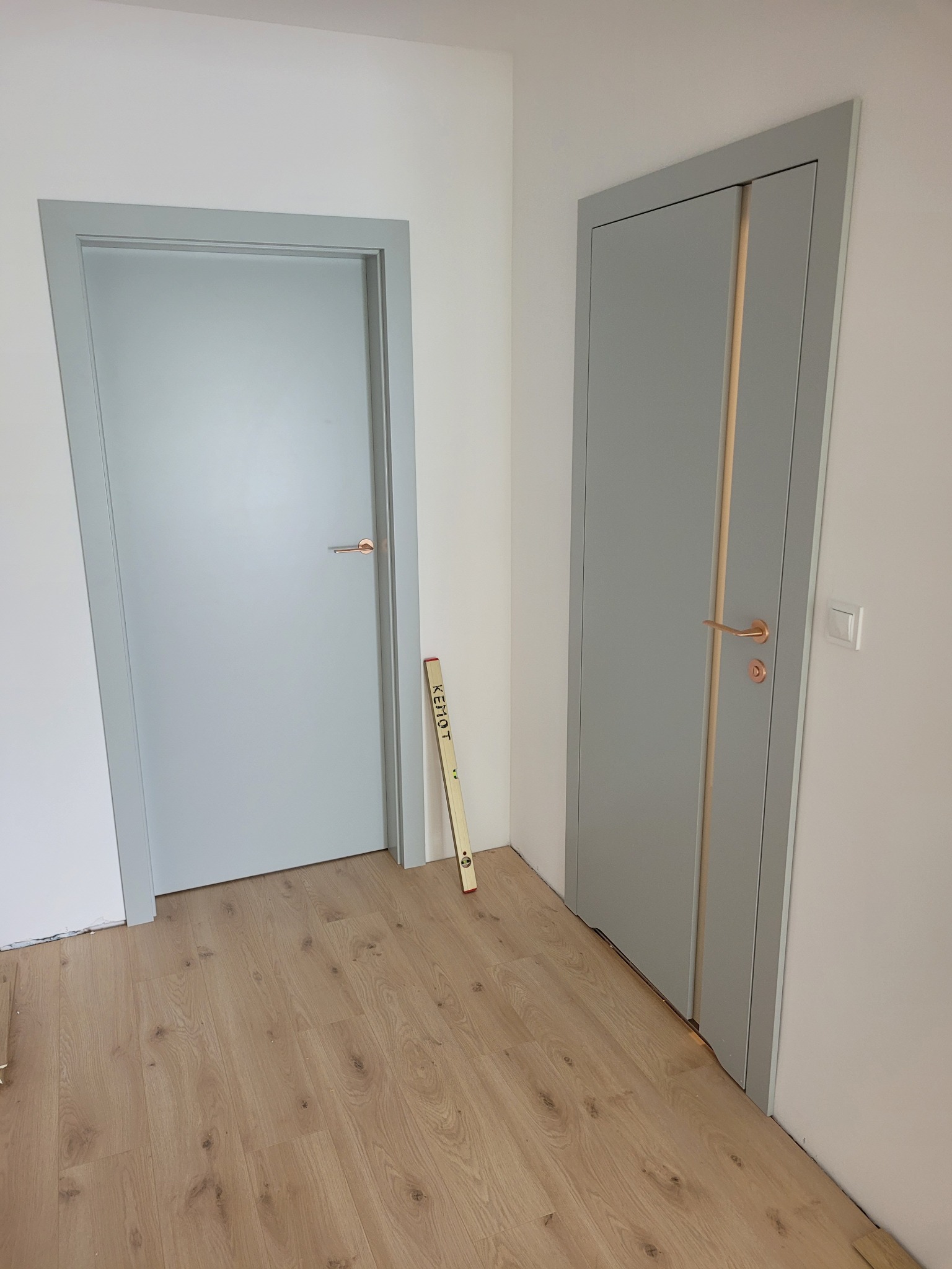 Dwa szare, nowoczesne skrzydła drzwiowe z miedzianymi klamkami, po montażu, na drewnianej podłodze. Widoczna poziomica oparta o ścianę.
