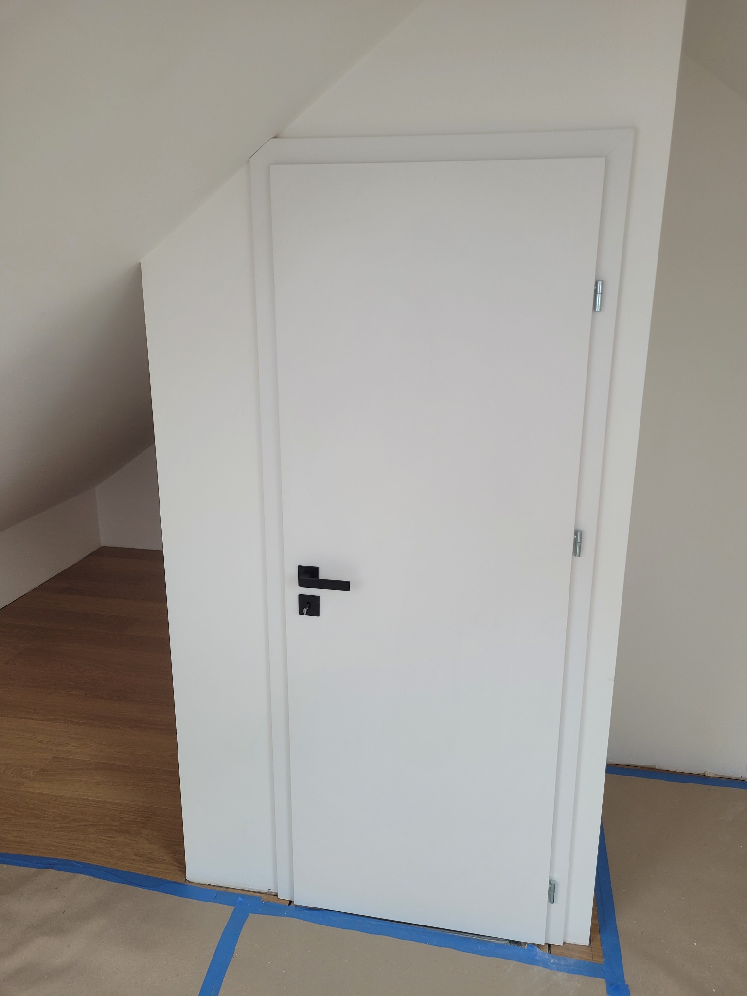 Białe, minimalistyczne drzwi z czarną klamką zamontowane w skosie poddasza. Widoczne zabezpieczenia podłogi taśmą malarską. Podłoga drewniana.