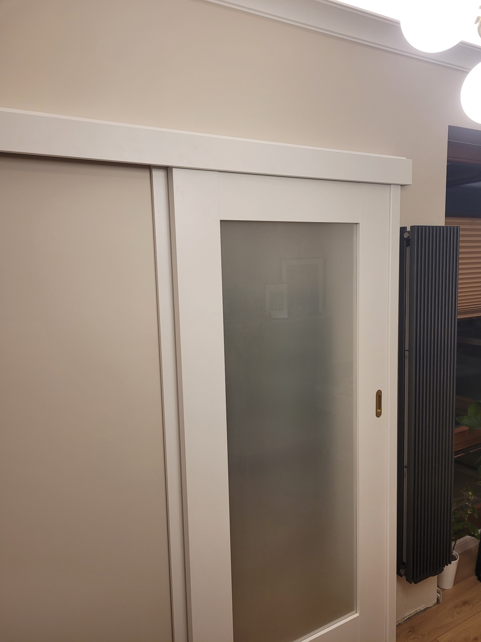 Białe przesuwne drzwi z matową szybą zamontowane w Trzyciąż. Widoczny fragment grzejnika i listwa przypodłogowa. Nowoczesny design, minimalistyczny styl.