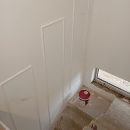 Usługi remontowe Walczak Damian - Świeżo pomalowana na biało ściana z dekoracyjnymi listwami, widok z góry na betonowe schody z wiadrem farby, okno w tle.