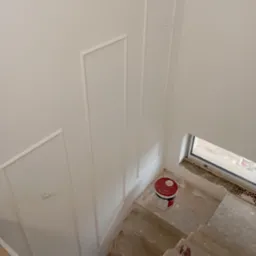Świeżo pomalowana na biało ściana z dekoracyjnymi listwami, widok z g&oacute;ry na betonowe schody z wiadrem farby, okno w tle.