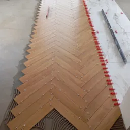 Układanie drewnopodobnych płytek podłogowych w jodełkę, z użyciem krzyżyków dystansowych i czerwonych klipsów poziomujących, widoczny fragment ściany i okna.