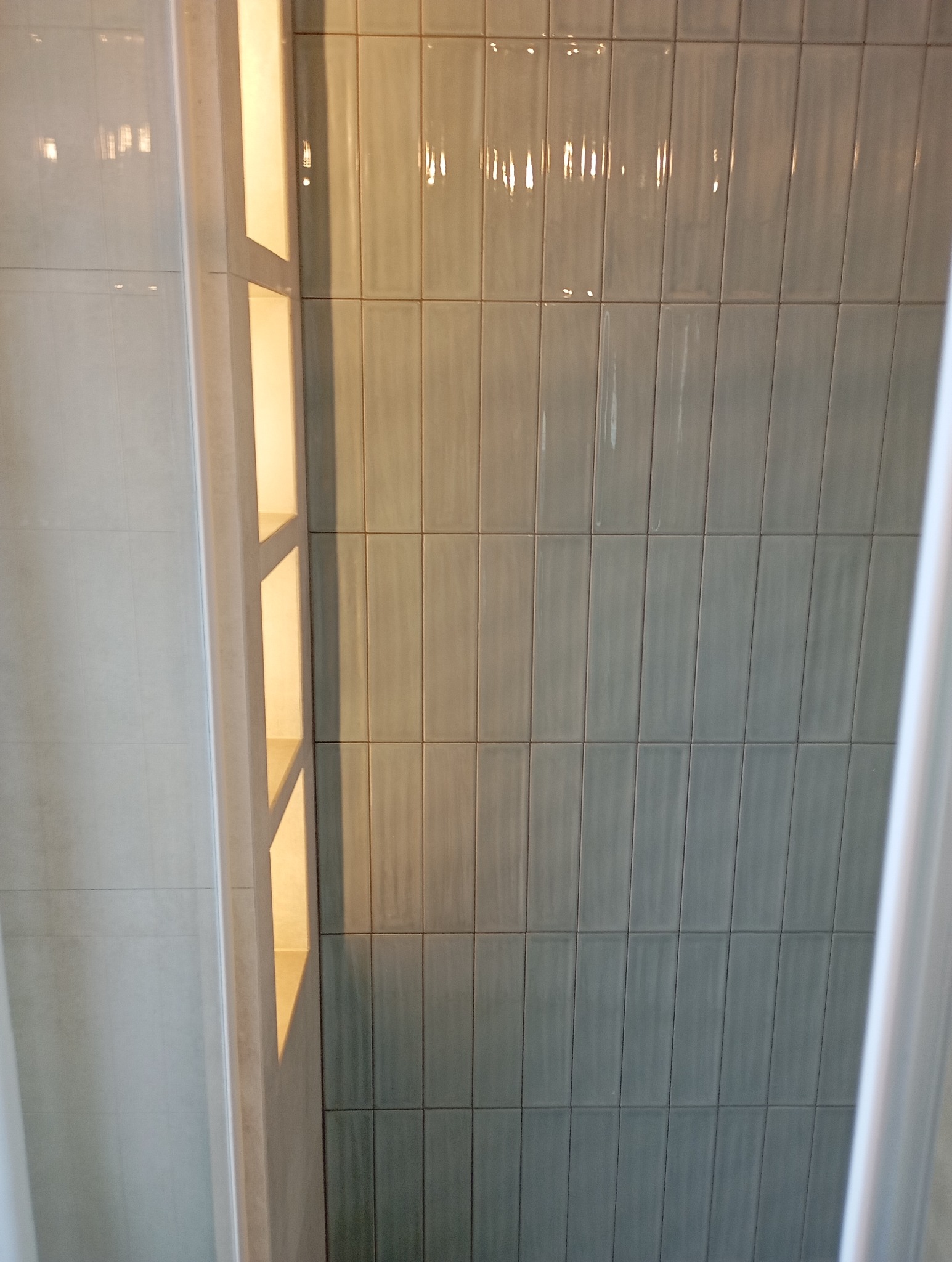 Błękitne, pionowo ułożone płytki w łazience, z widoczną wnęką z półkami po lewej stronie, oświetloną ciepłym światłem.