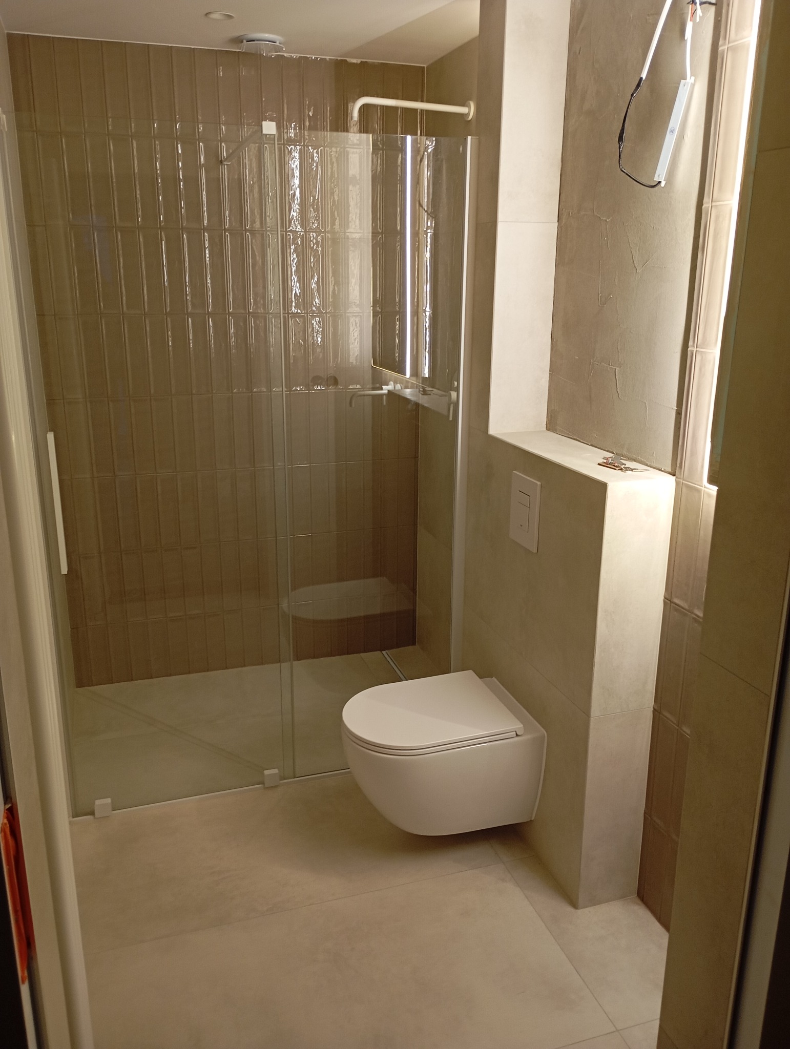 Nowoczesna łazienka z kabiną prysznicową z brązowymi kafelkami i podwieszaną toaletą, widoczne niedokończone prace przy oświetleniu lustra.