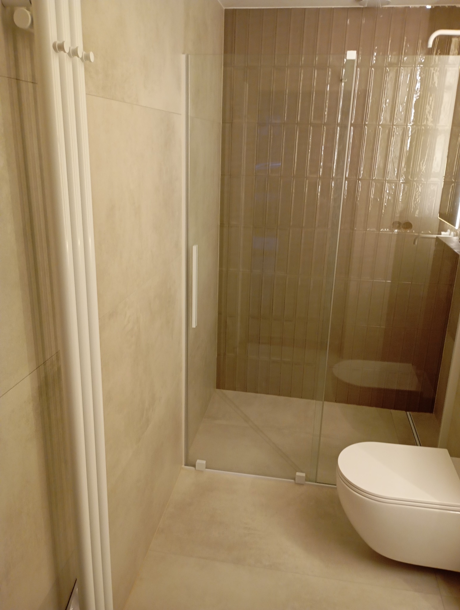 Nowoczesna, beżowa łazienka z kabiną prysznicową typu walk-in z długim, pionowym uchwytem, grzejnikiem drabinkowym i wiszącą miską WC.