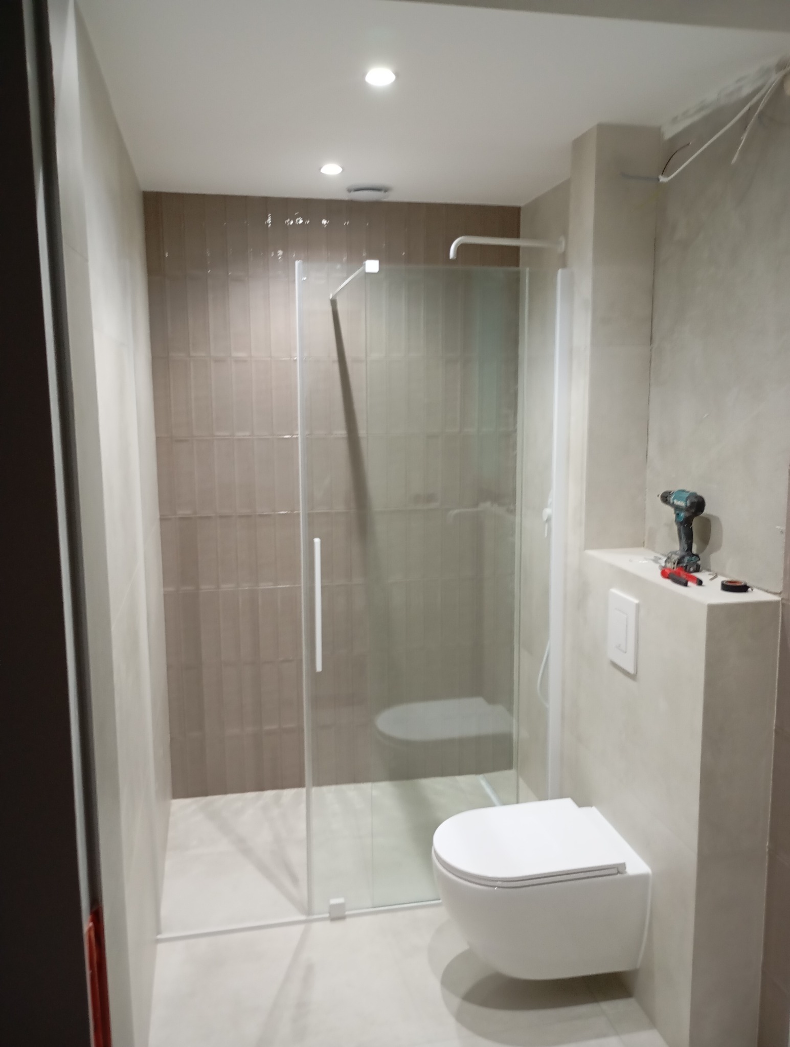 Nowoczesna łazienka z kabiną prysznicową typu walk-in, wiszącą toaletą i brązowymi płytkami na ścianie, w trakcie prac wykończeniowych, widoczna wiertarka i akcesoria na półce.