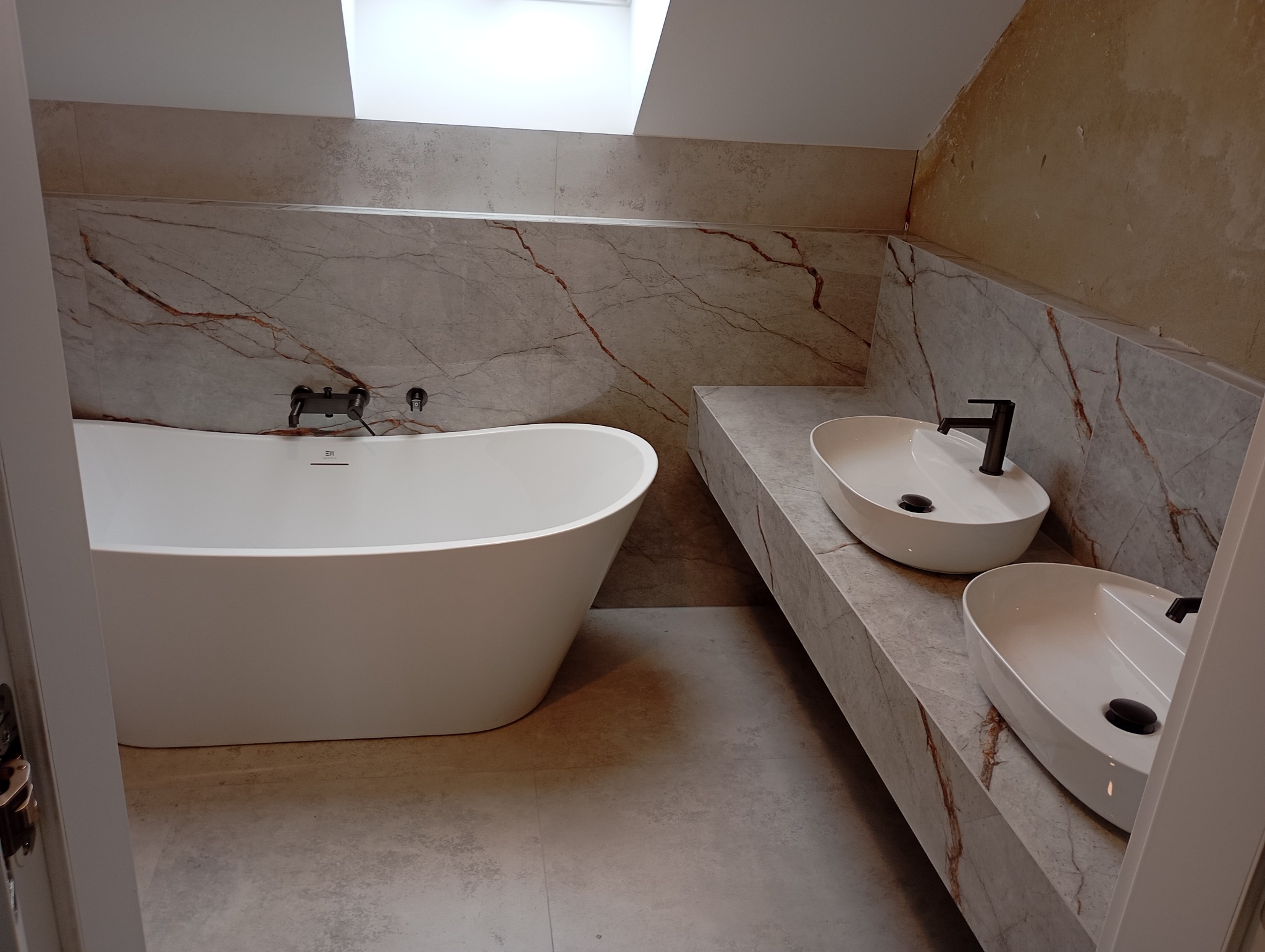 Nowoczesna łazienka z wolnostojącą białą wanną, dwoma owalnymi umywalkami nablatowymi z czarnymi bateriami na blacie z płytek imitujących marmur, oraz oknem dachowym.