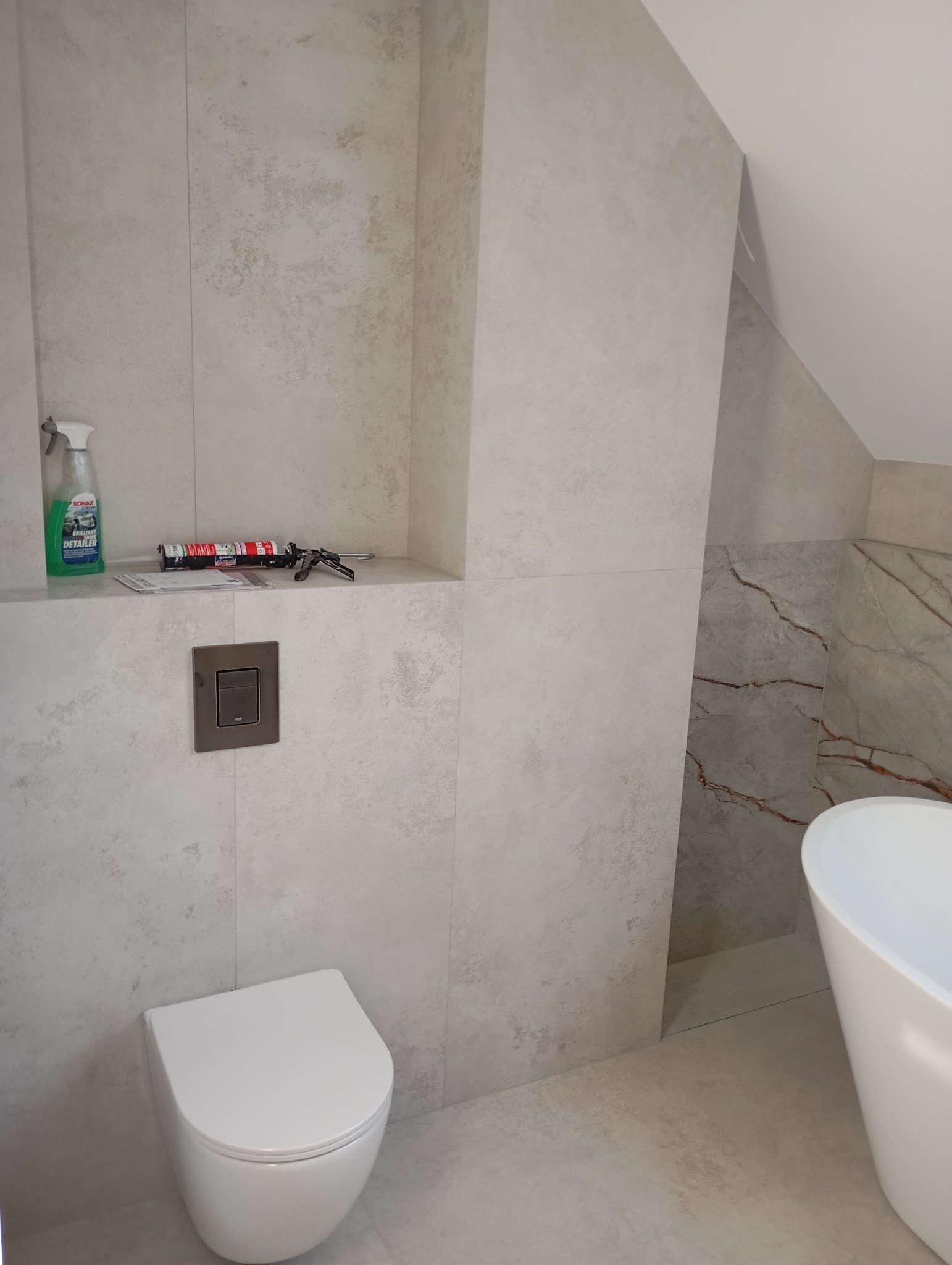 Nowoczesna łazienka z beżowymi płytkami imitującymi kamień, wiszącą toaletą, wolnostojącą wanną z marmurowym akcentem i półką z akcesoriami montażowymi oraz środkiem czyszczącym.