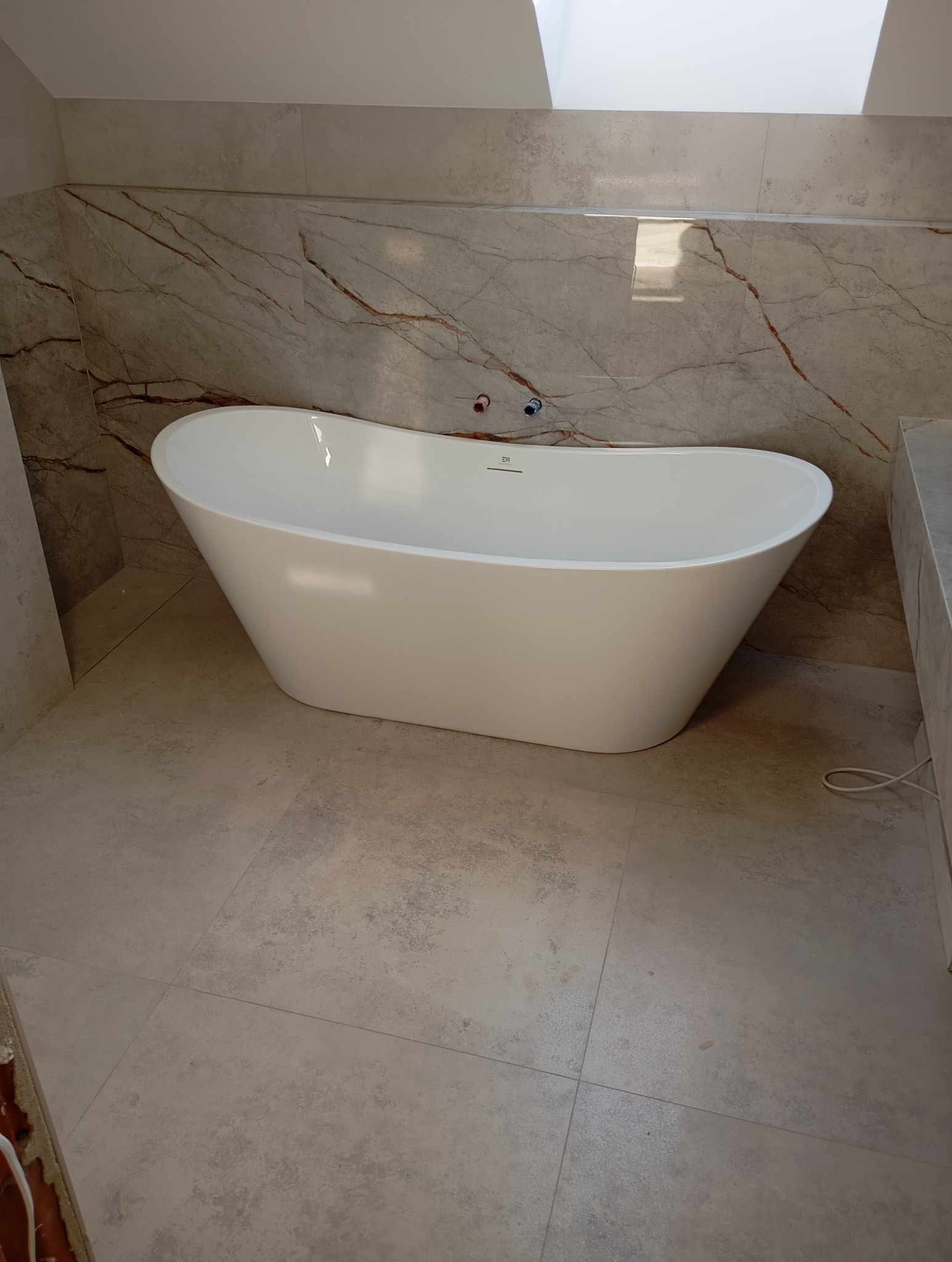 Wolnostojąca wanna o owalnym kształcie w łazience z marmurowymi płytkami na ścianach i podłodze. Widoczne instalacje wodne i okno dachowe.