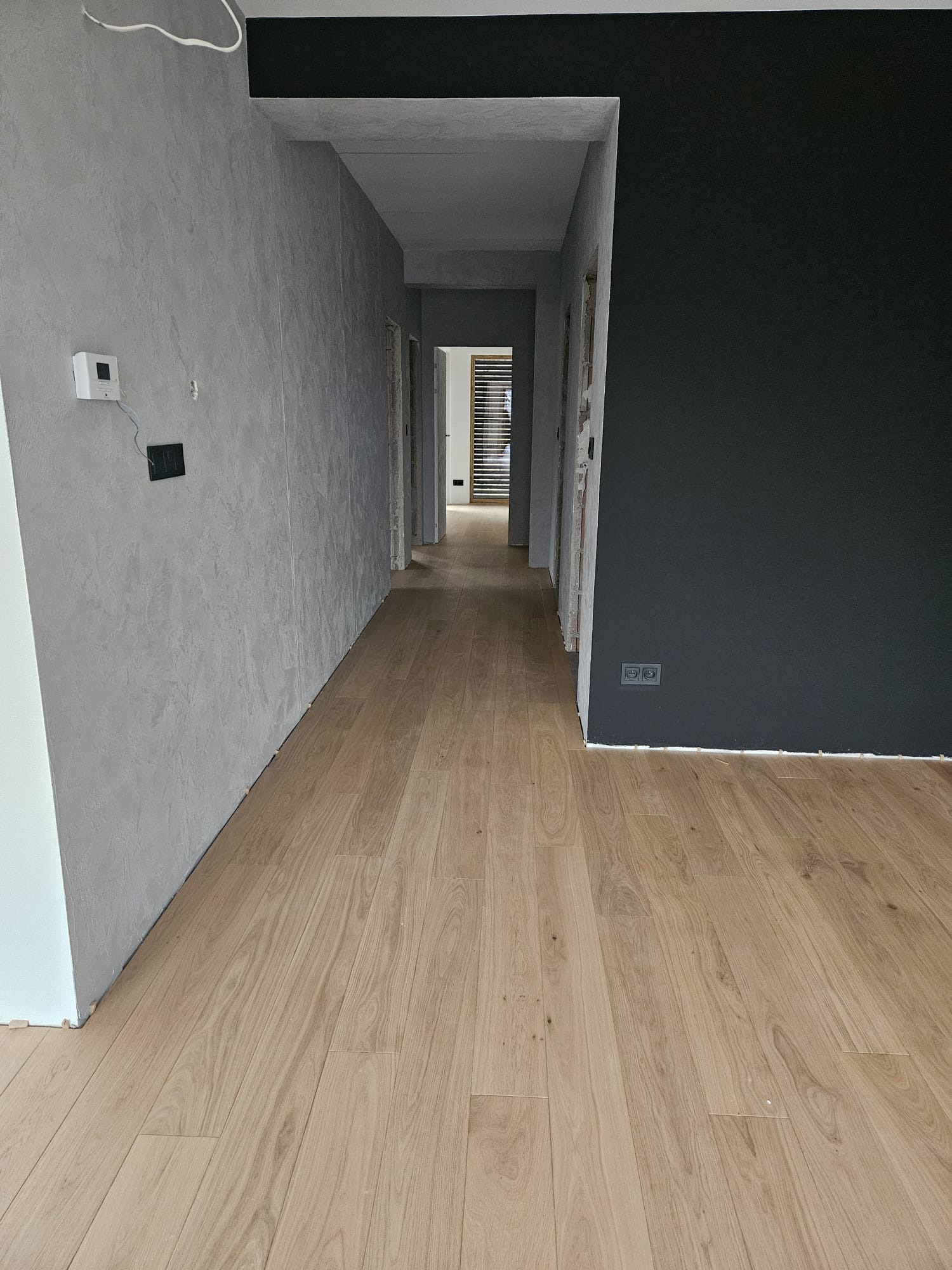 Wnętrze korytarza po remoncie: parkiet z jasnego drewna, ściana z szarą fakturą, fragment czarnej ściany z gniazdkami, widok w głąb prześwitu z drzwiami i żaluzjami.