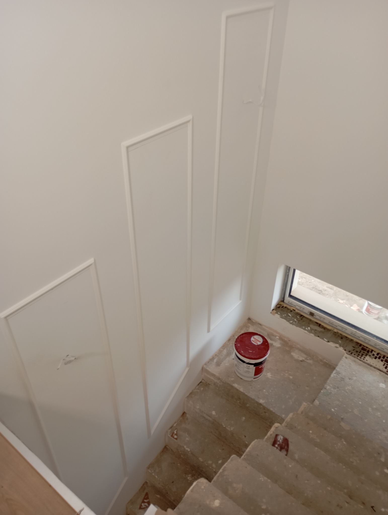 Świeżo pomalowana na biało ściana z dekoracyjnymi listwami, widok z góry na betonowe schody z wiadrem farby, okno w tle.