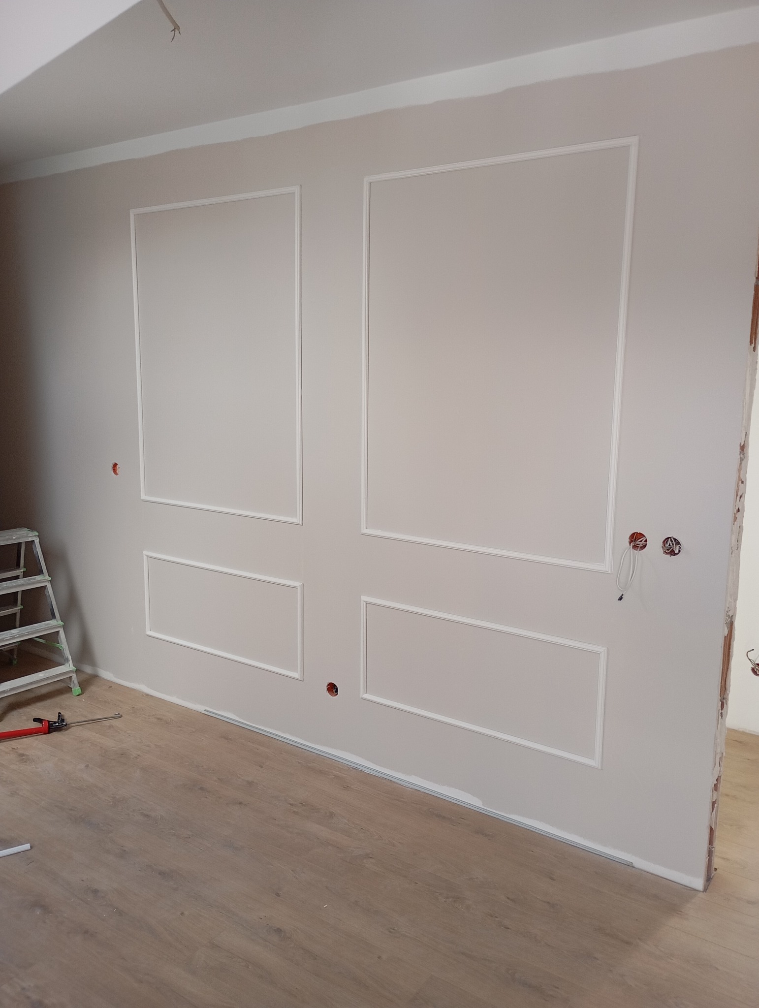 Ściana w trakcie dekorowania sztukaterią w postaci prostokątnych ram, widoczne narzędzia i drabina, surowy stan wykończenia podłogi i brakujące listwy przypodłogowe, gładź na ścianie w odcieniach...